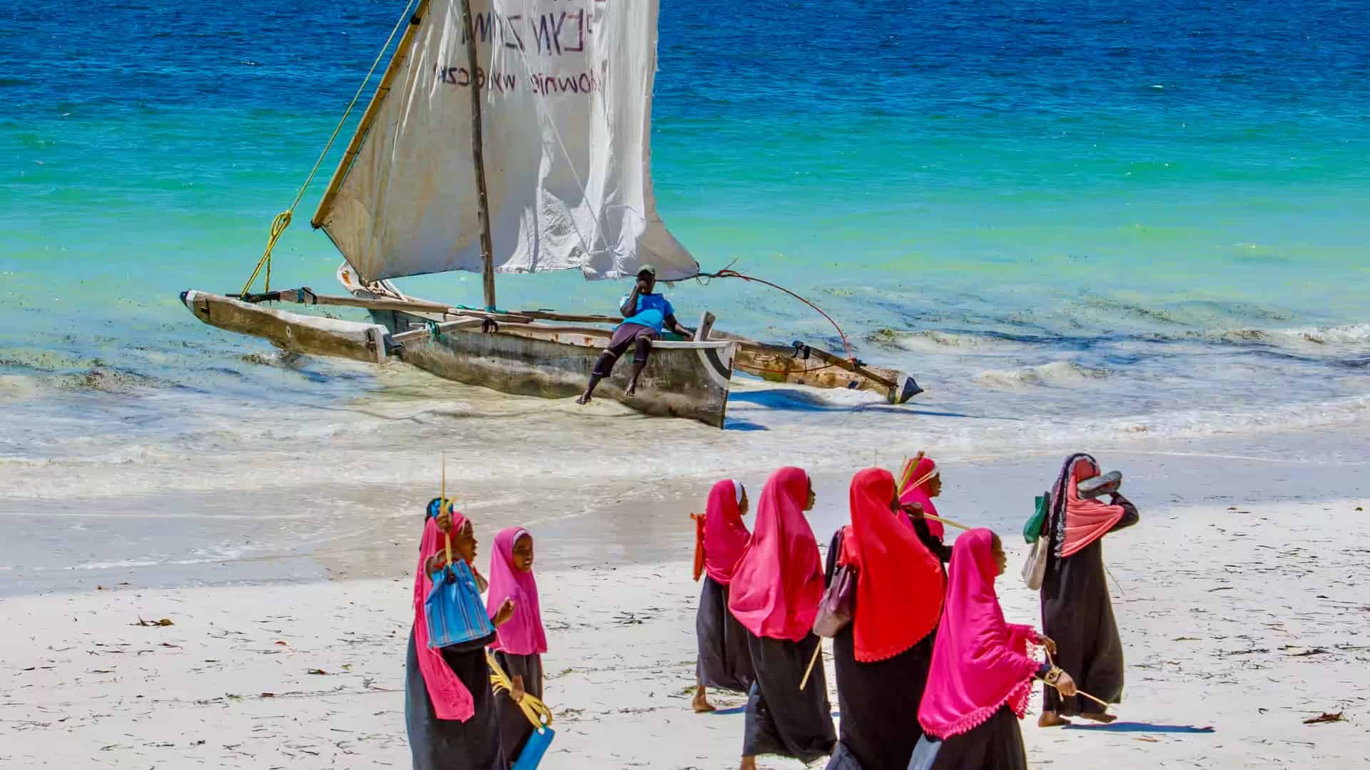 Tanzanian women walking along a pristine tropical beach in Zanzibar with a traditional wooden boat docked on the shore.