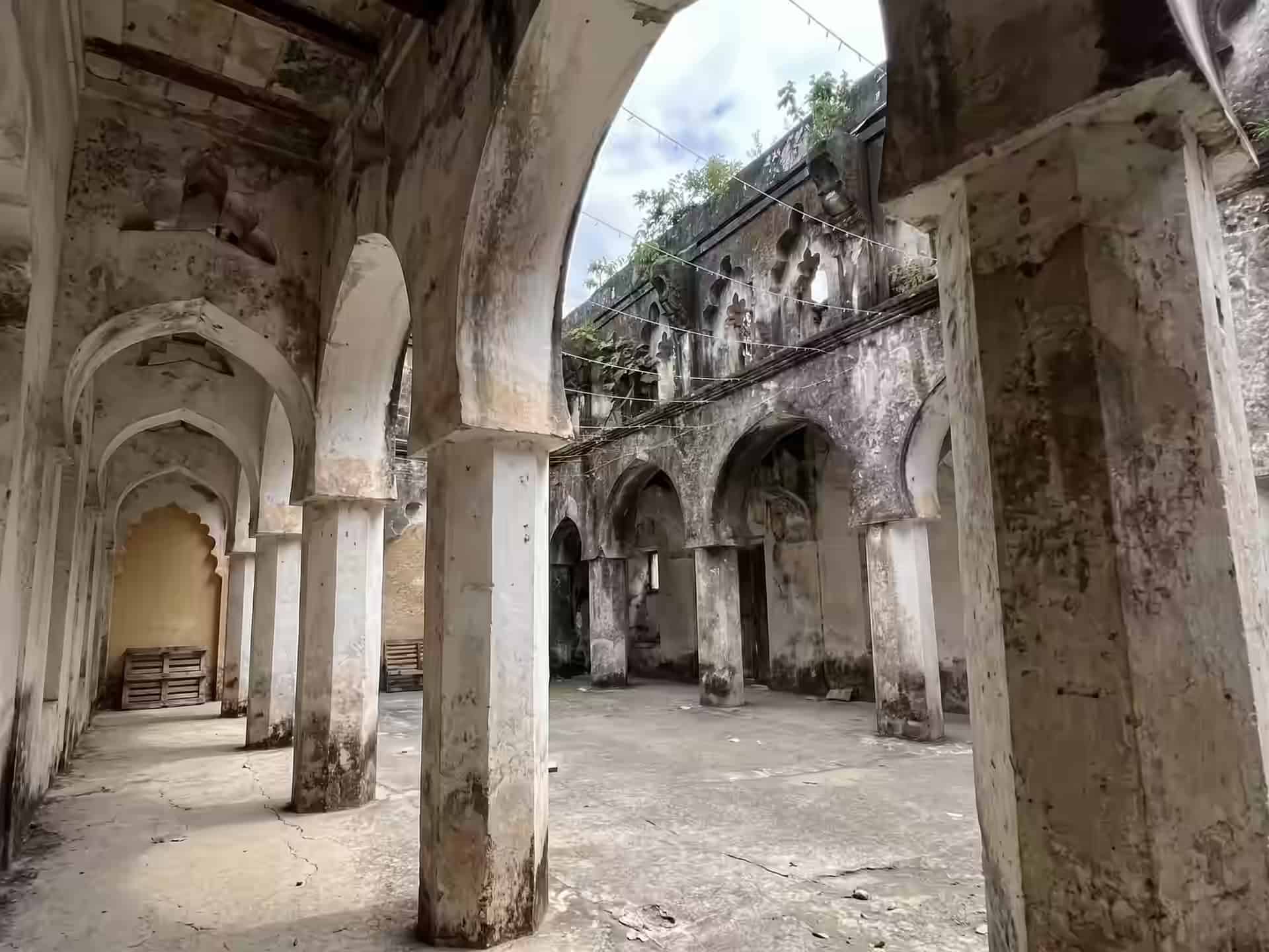 Verwitterte Arkaden aus Stein um einen offenen Hof in den Ruinen von Mbweni, auf den Mauerkronen wachsen Pflanzen.