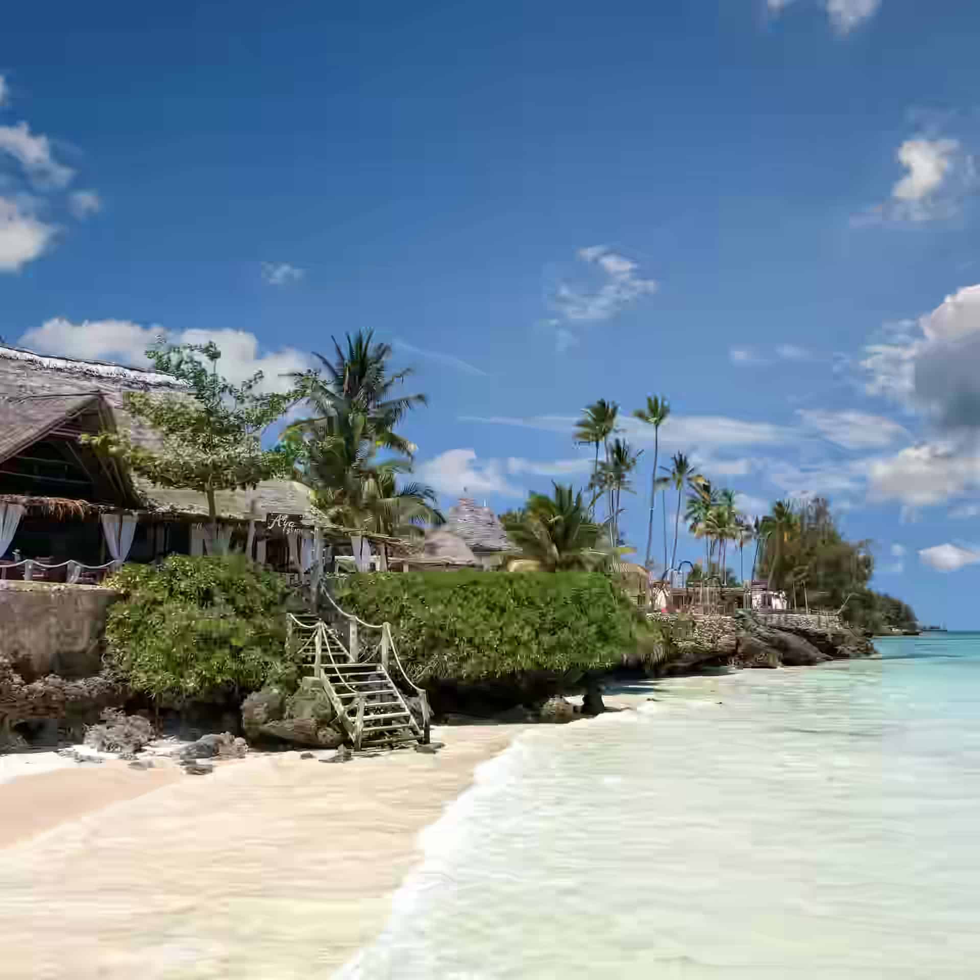 Ein Restaurant mit Strohdach auf einer felsigen Landzunge über dem Strand von Pongwe, mit Holzstufen hinunter zum weißen Sand, Palmen und türkisfarbenem Wasser.