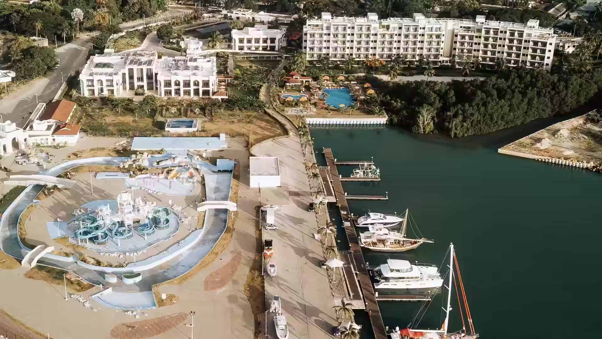 Luftbild eines Jachthafens mit Jachten und Segelbooten an einem Steg, neben einem Hotel am Meer, Pools und einem Wasserpark.